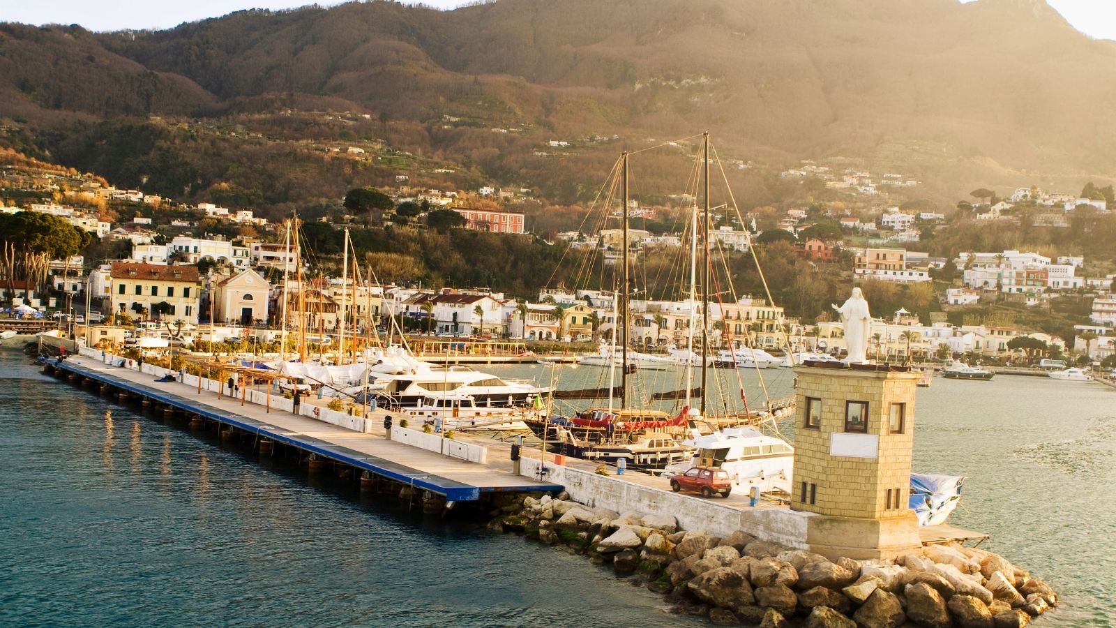 Porto di Casamicciola Terme a Ischia: barche e velieri ormeggiati nella luce del tramonto con le colline verdi sullo sfondo